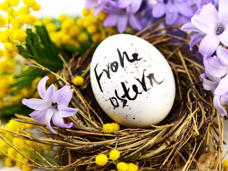 In einem kleinen Nest aus dünnen Ästen ist ein Ei drin, wo frohe Ostern drauf geschrieben ist. Im Hintergrund sind viele bunte Blumen.