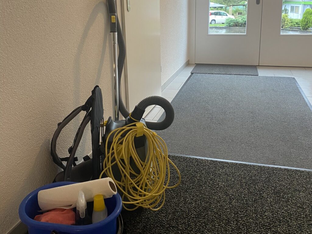 Ein Hauseingang, mit verschiedenen Teppichen am Boden es ist ein Staubsauger und eine Eimer mit Reinigungsutensilien ersichtlich
